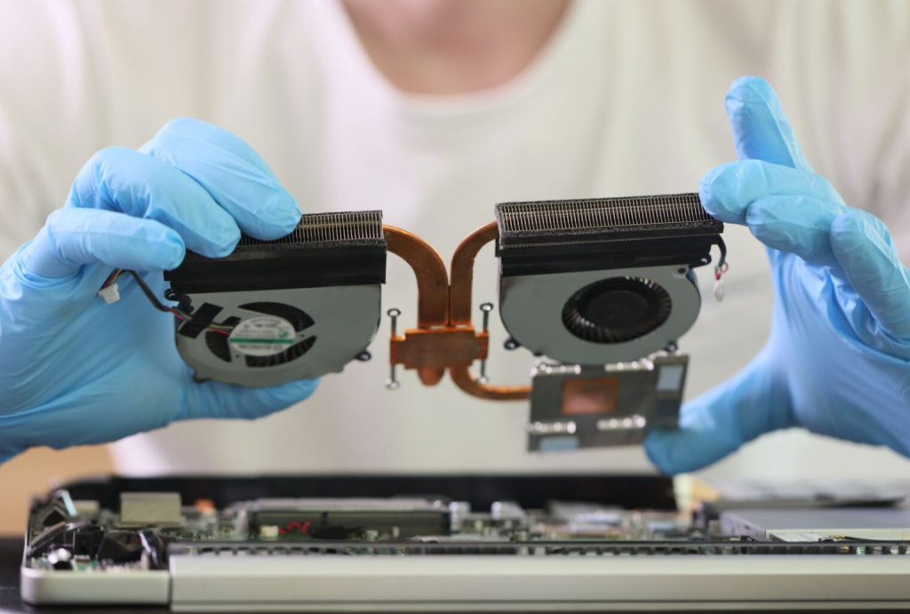 A technician in blue gloves holding a dual-fan laptop cooling assembly with copper heat pipes during a hardware repair.